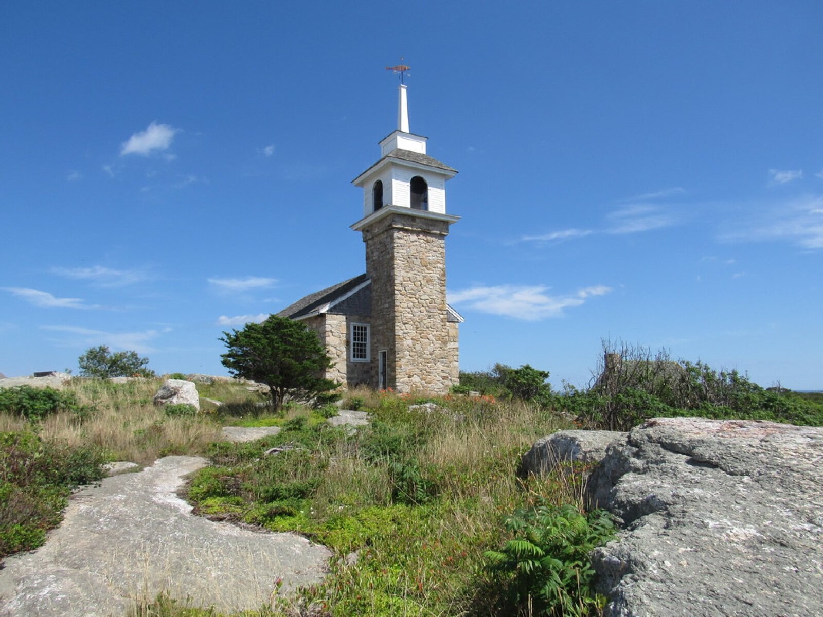 The stone Gosport Chapel with a white bell tower on a rocky, grassy rise on Star Island under a blue sky