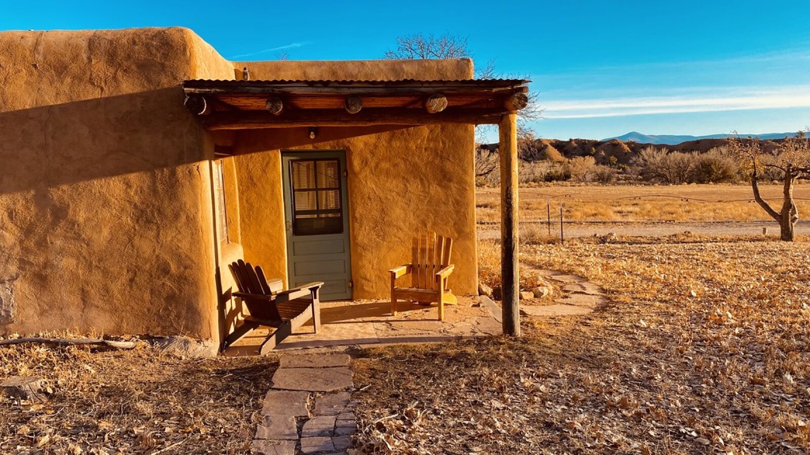 A small ochre adobe casita with a wooden porch and two chairs, set among dry grass and cottonwood leaves under a blue sky at Ghost Ranch