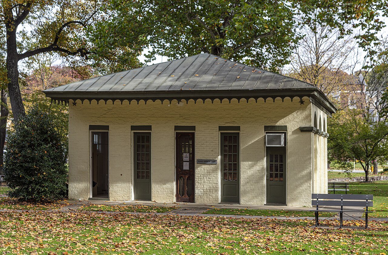 A small cream-painted brick building with a metal hipped roof and green doors, the 1845 park office, among autumn trees in Berkeley Springs State Park