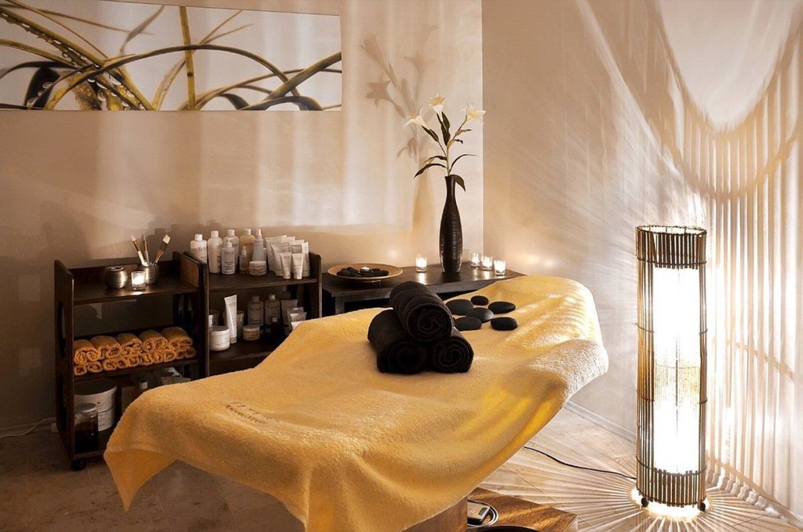 A softly lit spa treatment room with a massage table, rolled towels, a lit lantern and a single lily