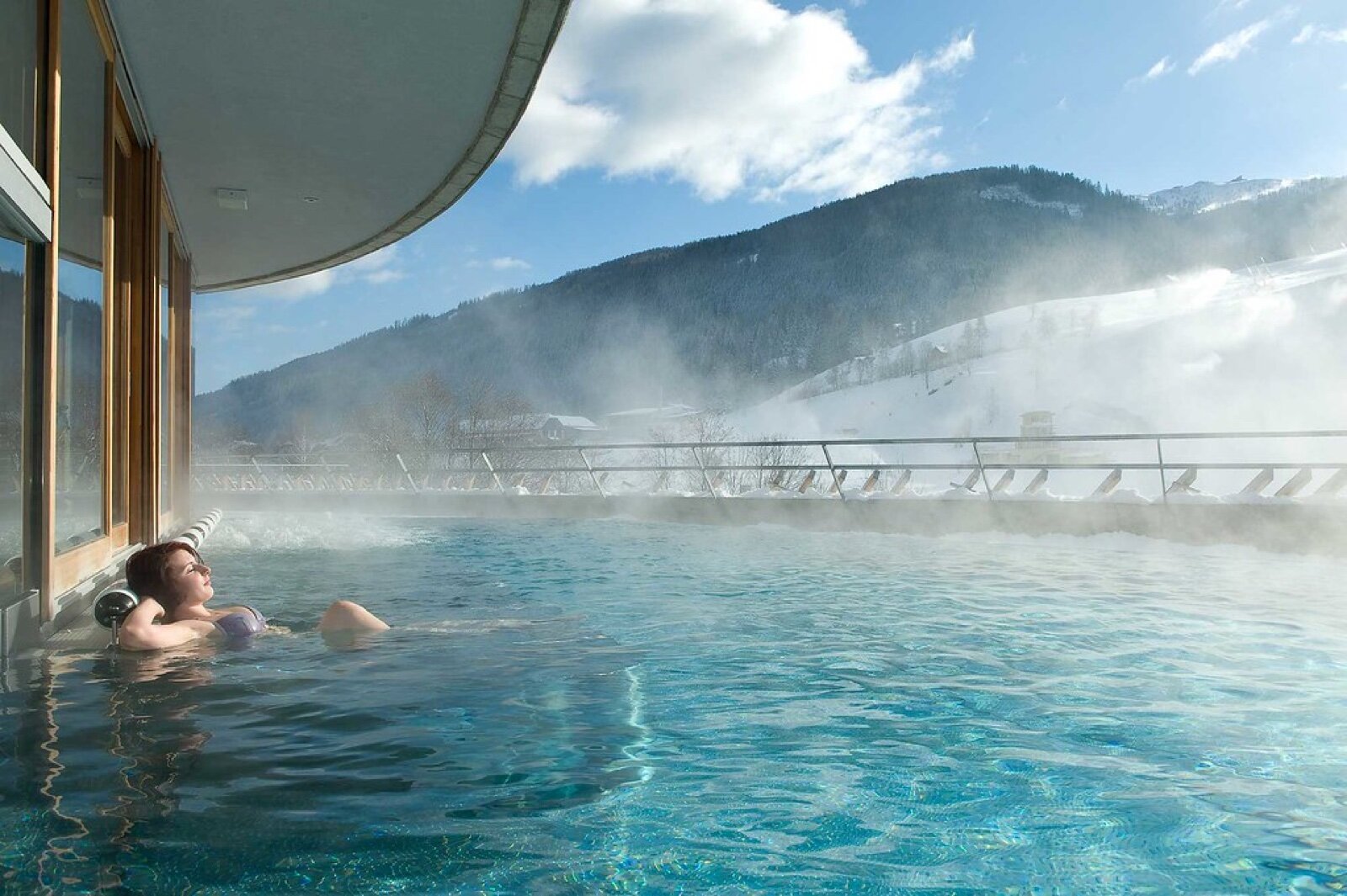 An outdoor thermal spa pool with steam rising against a snowy alpine mountain backdrop